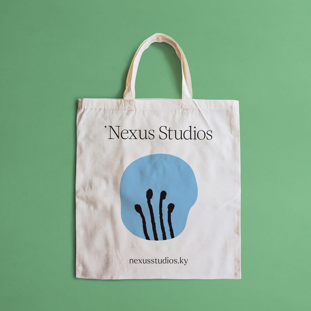 A canvas tote bag from Nexus Studios placed on a soft green background. The tote bag is white and displays the "Nexus Studios" logo in black serif text at the top. Below the logo, there is a distinctive graphic consisting of a light blue circle with black stylized matchstick figures inside it, suggesting a creative or artistic theme. The website URL "nexusstudios.ky" is printed in small black letters at the bottom of the bag. The overall design of the tote bag is clean and visually engaging, reflecting the brand’s creative and modern aesthetic.