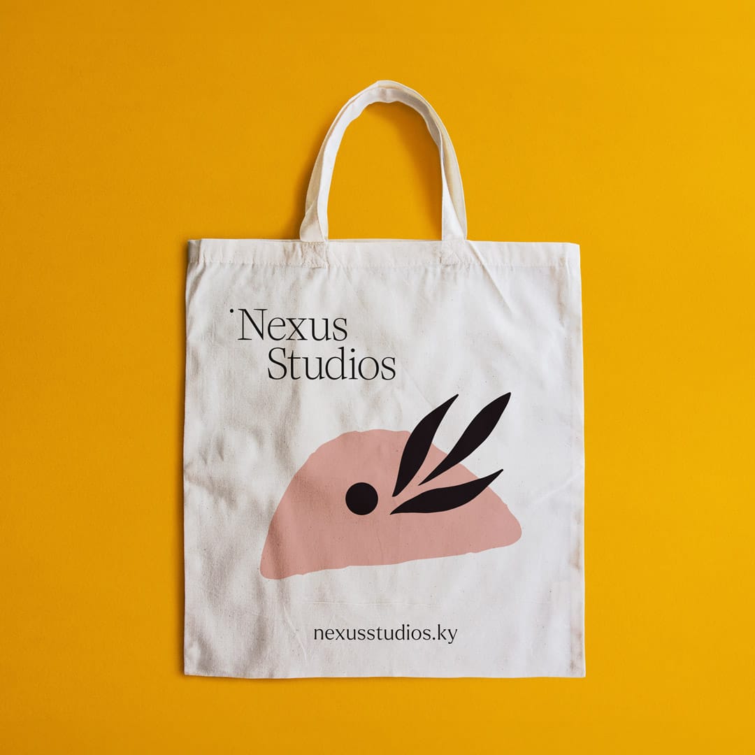 A canvas tote bag from Nexus Studios placed on a vivid yellow background. The bag is predominantly white with the Nexus Studios logo in black serif text centred near the top. Below the logo, there's a large graphic of a stylized black leaf with black details, creating an eye-catching visual element. The website URL "nexusstudios.ky" is printed in small black letters at the bottom of the bag. The design is simple yet stylish, effectively conveying the brand’s modern aesthetic.