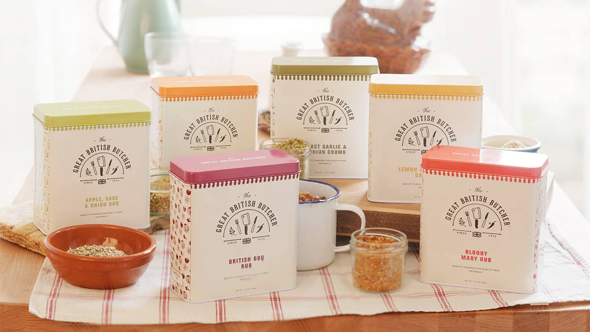 Each of the 6 Great British Butcher product tins situated in a traditional rustic setting on a wooden farmhouse table. The tins at the front are placed on top of a checked tea towel, and the ones at the back on a chunky wooden chopping board. There is an enamel mug and a couple of ceramic bowls containing rubs and crumbs. In the background is a vintage jug and an old school ceramic hen - used to store eggs.