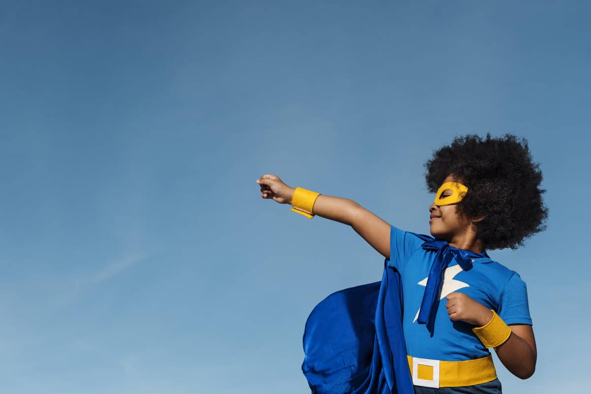 A child of colour with afro hair and a blue super hero costume with a lightning bolt on their chest. They are also wearing a dark blue cape and have a bright yellow eye mask, belt and wrist bands. The photo is taken against a blue sky. The child has their right hand up in a fist pointing into the distance as if they were ready to take action or fly.