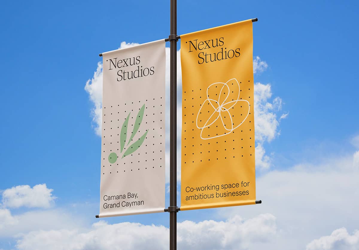 Two vertical banners mounted on a single flagpole. The left banner is white with the logo of "Nexus Studios," featuring a stylised green leaf and black dot pattern along with the text "Camana Bay, Grand Cayman" in a serif font. The right banner is golden yellow with a large, abstract, white outline of flower drawing that similar to an atom model, surrounded by a dotted pattern. It includes the text "Nexus Studios" at the top and "Co-working space for ambitious businesses" at the bottom, both in a serif font. The banners are depicted fluttering slightly, suggesting a gentle breeze.