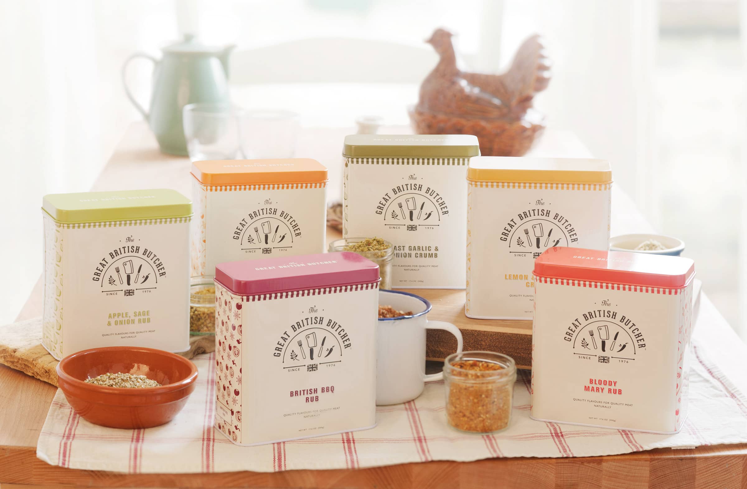 Each of the 6 Great British Butcher product tins situated in a traditional rustic setting on a wooden farmhouse table. The tins at the front are placed on top of a checked tea towel, and the ones at the back on a chunky wooden chopping board. There is an enamel mug and a couple of ceramic bowls containing rubs and crumbs. In the background is a vintage jug and an old school ceramic hen - used to store eggs.