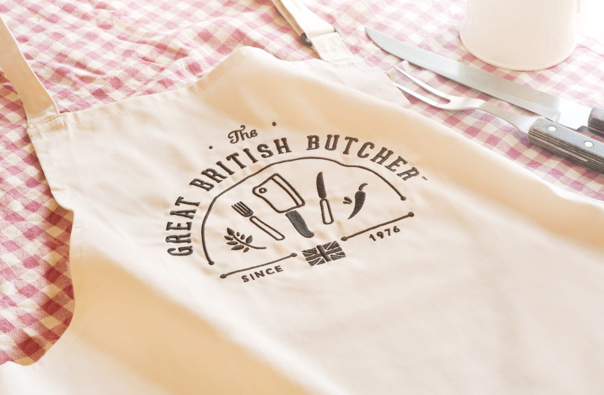 A cream apron with the Great British logo embroidered on to the front lying on a check tablecloth